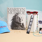 Collection of items including a book titled 'Marilyn Monroe: A Portrait', sunglasses, and a blue cap on a beige surface.