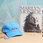 Blue cap with red text, 'Marilyn Monroe' book, and bracelet on a blue and beige background.