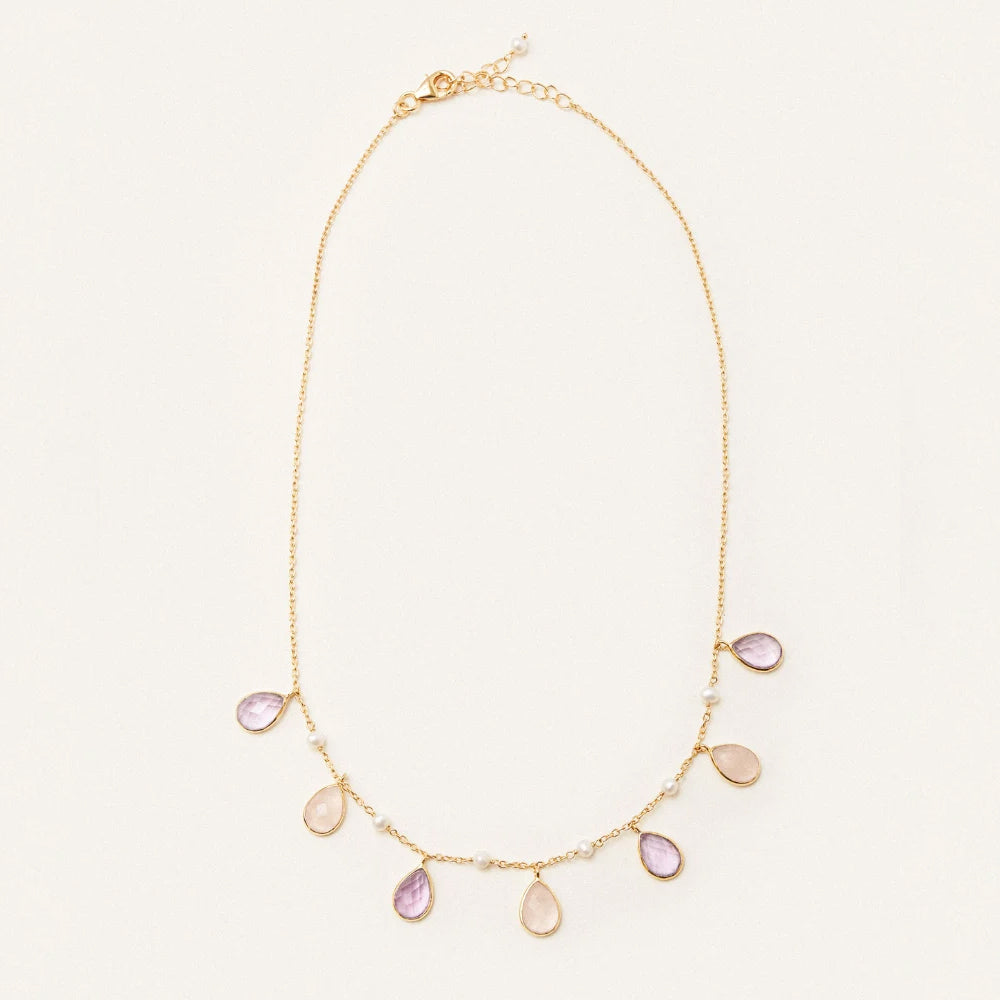 A delicate gold chain necklace featuring seven teardrop shaped pale pink gemstone pendants and small pearl like beads, displayed on a clean off white background.