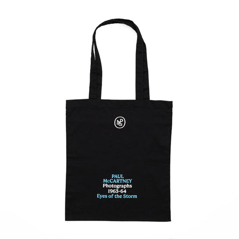 Black tote bag printed with the exhibition title 'Paul McCartney Photographs 1963-64 Eyes of the Storm' in blue and white. 