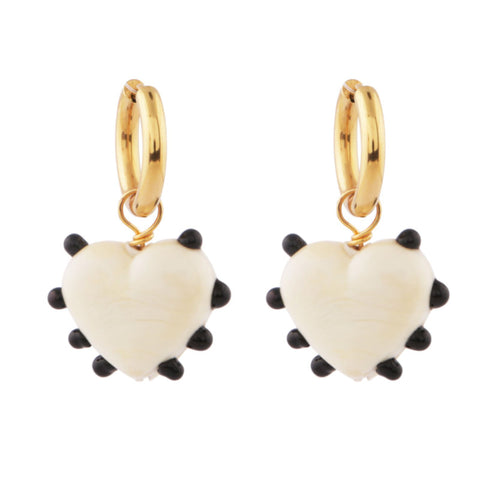 A pair of ivory glass heart earrings with black dots hanging from gold hoops.