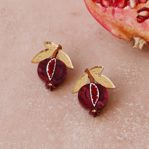 A pair of red and gold 3D acrylic pomegranate earrings in front of a pomegranate fruit.