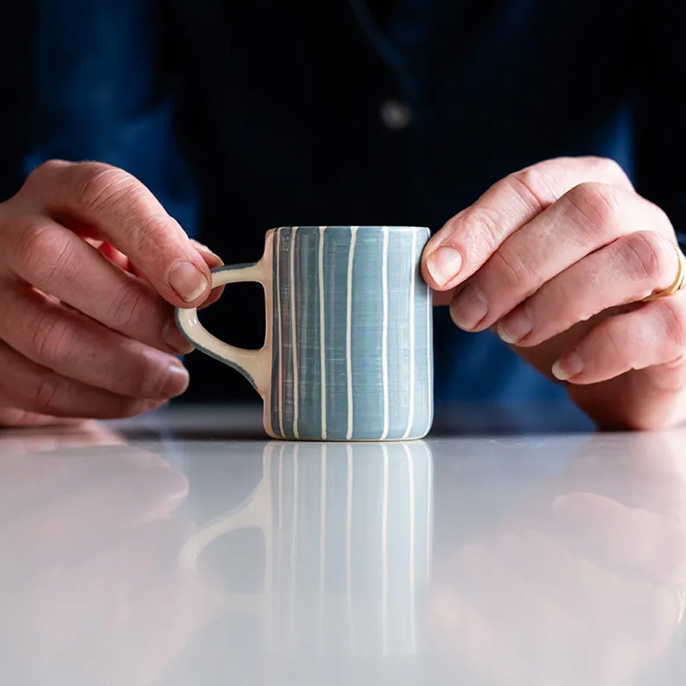 A pair of hands holds a small ceramic cup with handle, decorated with painted grey and white stripes.