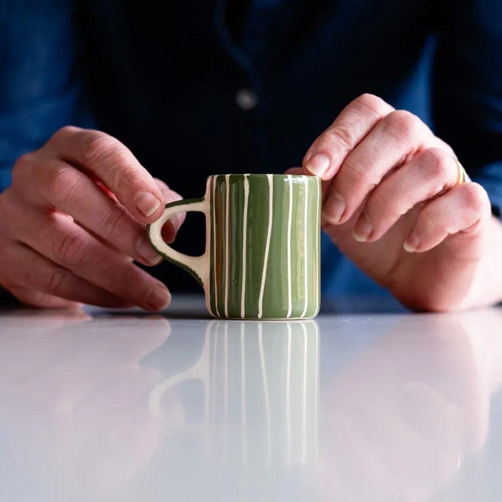 A pair of hands hold a ceramic cup with a handle, painted in green and white stripes.