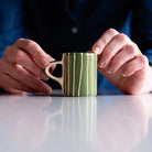 A pair of hands hold a ceramic cup with a handle, painted in green and white stripes.