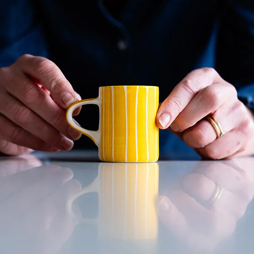 A pair of hands holds a small ceramic cup with handle, decorated with painted stripes in yellow and white.