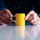 A pair of hands holds a small ceramic cup with handle, decorated with painted stripes in yellow and white.