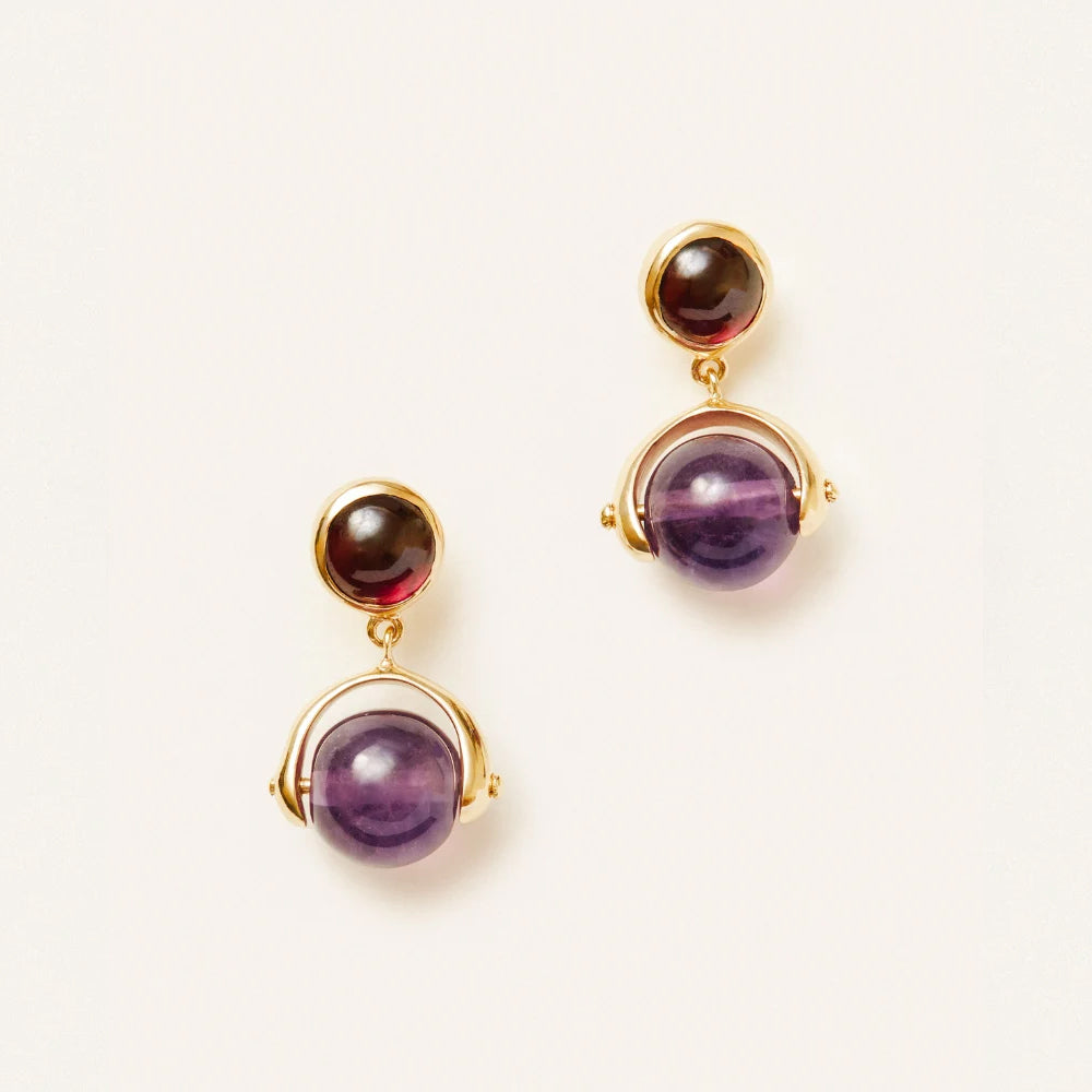 A pair of gold drop earrings featuring a small round dark stone at the top and a larger round purple stone at the bottom, set against a plain off white background.