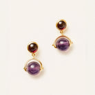 A pair of gold drop earrings featuring a small round dark stone at the top and a larger round purple stone at the bottom, set against a plain off-white background.