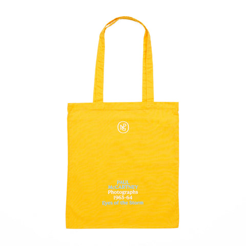 Yellow tote bag printed with the exhibition title 'Paul McCartney Photographs 1963-64 Eyes of the Storm' in blue and white. 