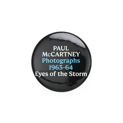 Small black round badge with the exhibition title text 'Paul McCartney Photographs 1963-64 Eyes of the Storm' in white and blue.