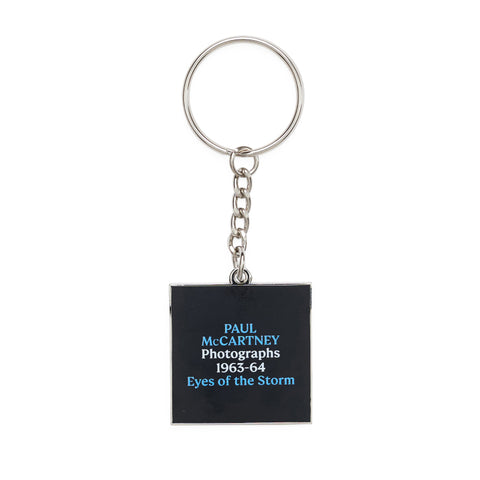 Square black enamel keyring with the exhibition text 'Paul McCartney Photographs 1963-64 Eyes of the Storm' printed in blue and white.