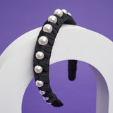 A black headband with fabric ruffles and large pearls balances on a white arch.