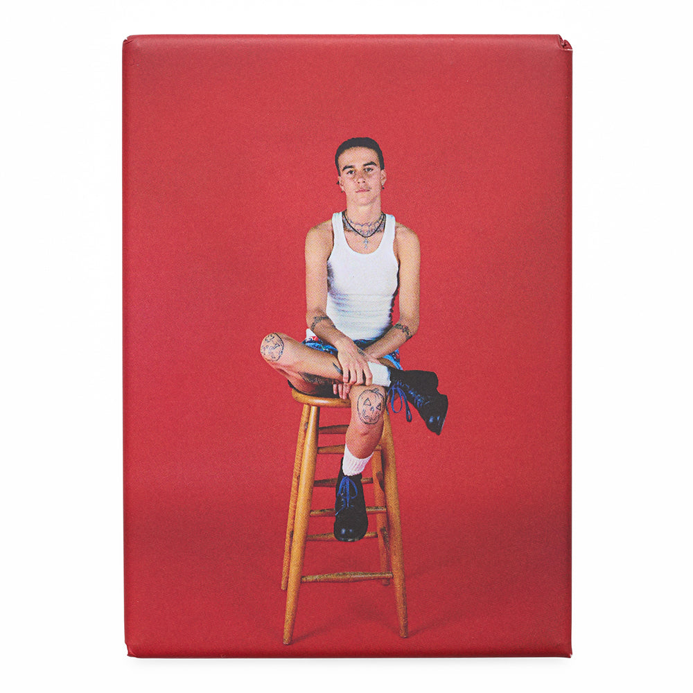 Magnet with a photograph of a person sitting on a wooden stool against a red backdrop. 