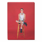 Magnet with a photograph of a person sitting on a wooden stool against a red backdrop. 
