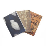 A collection of 3 sketchbooks. One is ochre and has black floral designs, one is beige and tan with leaf designs and the third is blue with white cloud-like motifs.
