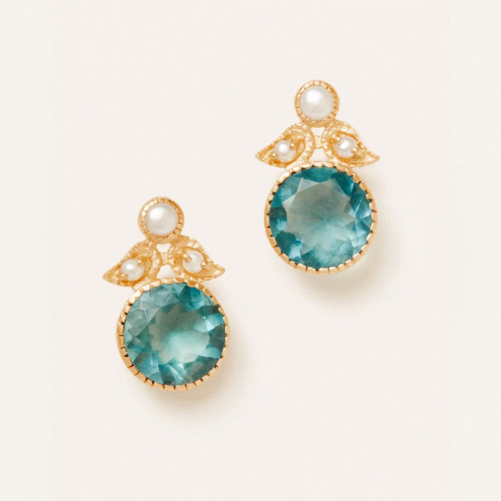 A pair of gold drop earrings featuring large round faceted teal blue gemstones, small white pearl accents, and delicate gold leaf like details, set against a clean off white background.