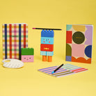 Colorful stationery items including notebooks, a pencil holder, and a paint palette on a yellow background.