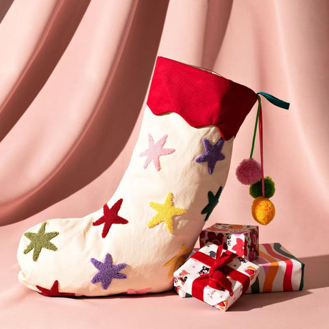 A white fabric stocking with multicoloured stars on it and colourful pompoms. All against a pink background with three small presents resting next to it.