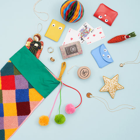 A multicoloured chequered fabric stocking with colourful pompoms and multiple gifts falling out of the top including a yoyo, purse, playing cards and decorative angel.