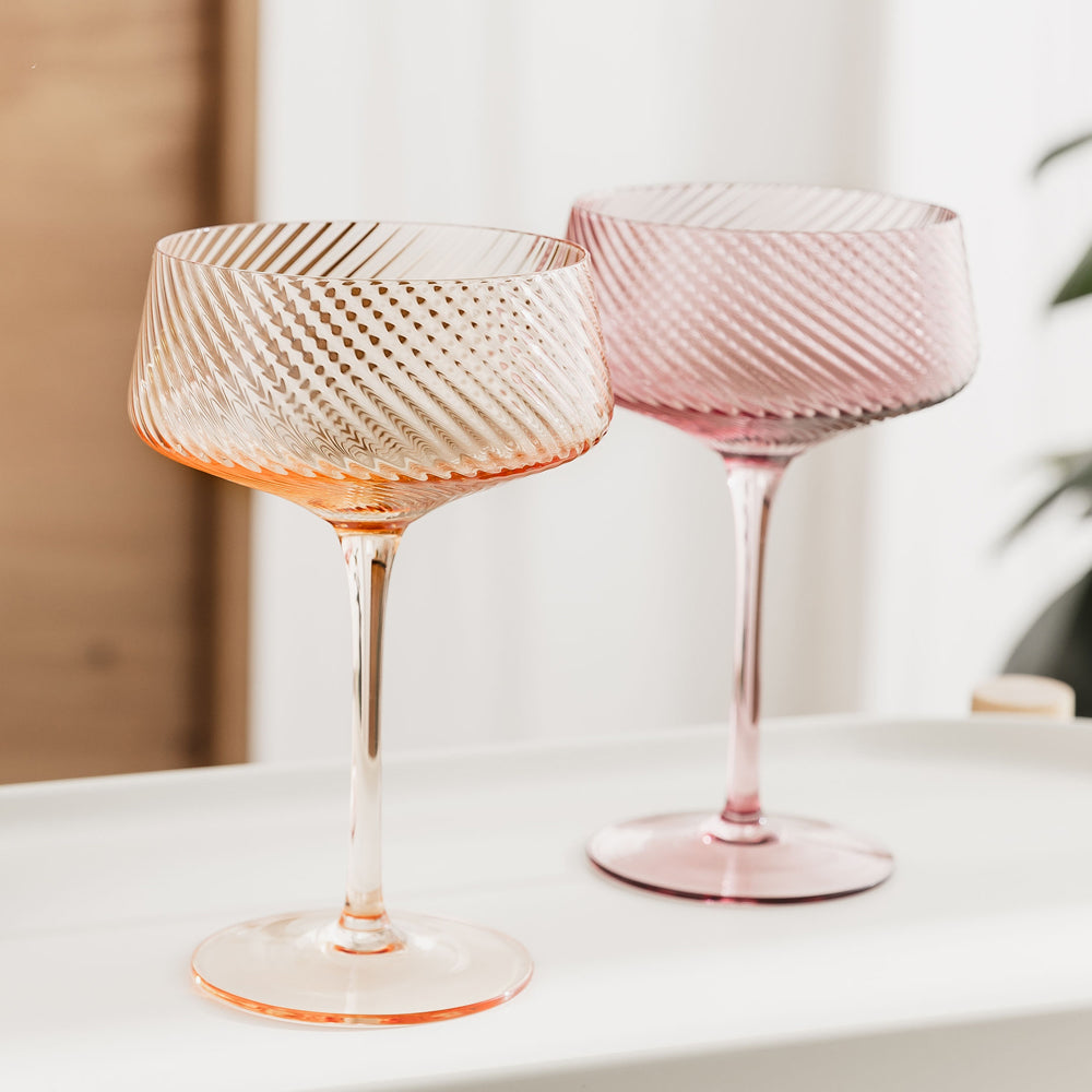 Two stemmed cocktail glasses with textured, diamond patterned bowls, one in translucent pink and one in translucent orange, displayed on a white surface.
