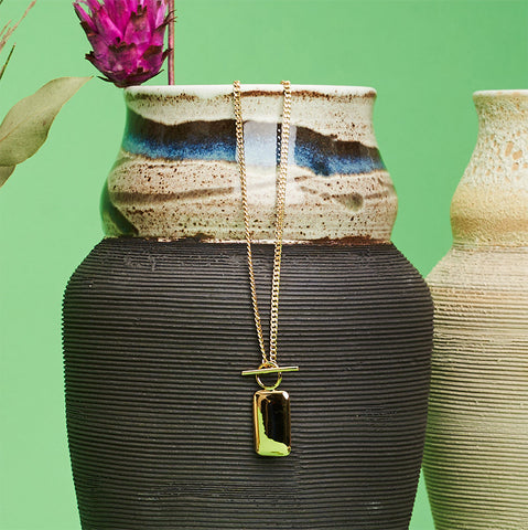 Gold rectangular pendant on a chain hanging from a ceramic vase. 