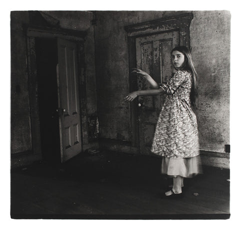 Photographic postcard of a woman holding out her arms in front of an open door. 