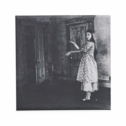Square magnet featuring a black and white photograph of a girl in a dress in front of a door. 