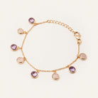 A delicate gold chain bracelet with seven round bezel-set charms, including four purple amethyst stones and three pale pink stones, displayed on a clean off-white background.